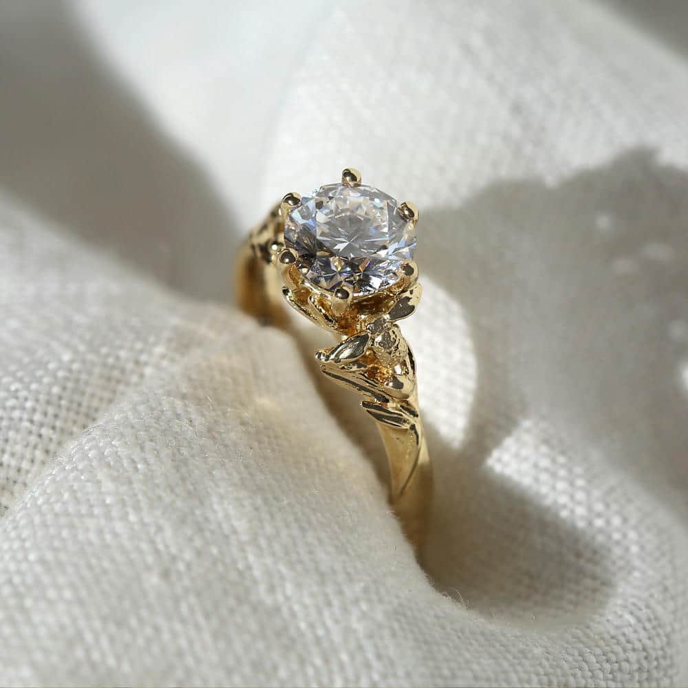 Close up of an 18ct yellow gold solitaire diamond ring with gold bee and floral details on white fabric backdrop.