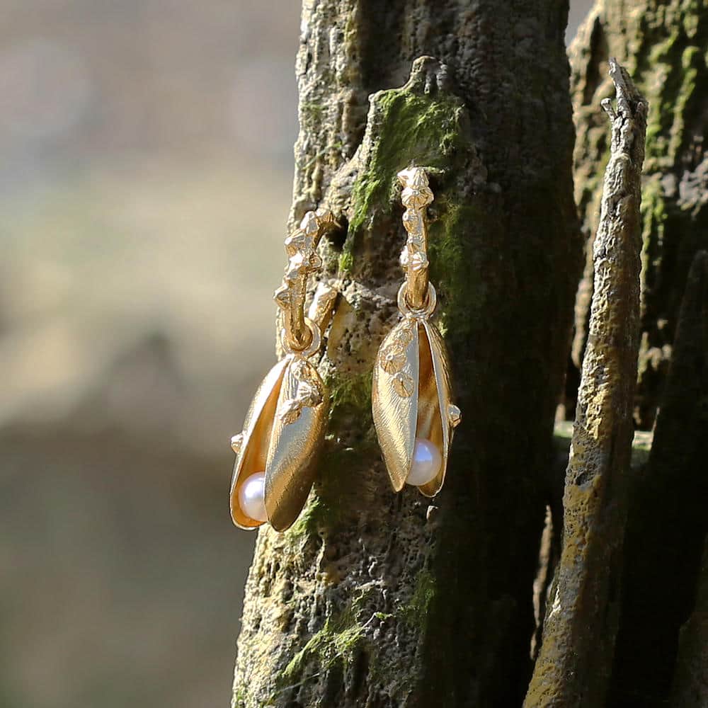 Gold drop earrings designed as delicate mussel shells with tiny engraved details, each holding a freshwater pearl, photographed hanging against textured driftwood.