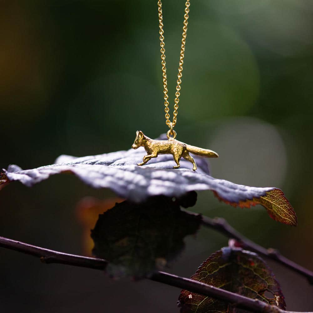 Prowling Fox Necklace - a finely detailed gold fox pendant on a delicate chain, displayed resting on an autumn leaf