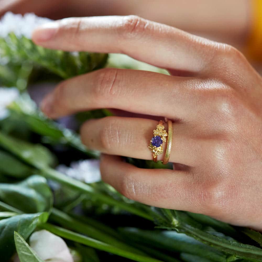 Hand wearing a gold ring set with a deep blue sapphire, styled with floral detailing, photographed among fresh green leaves and flowers