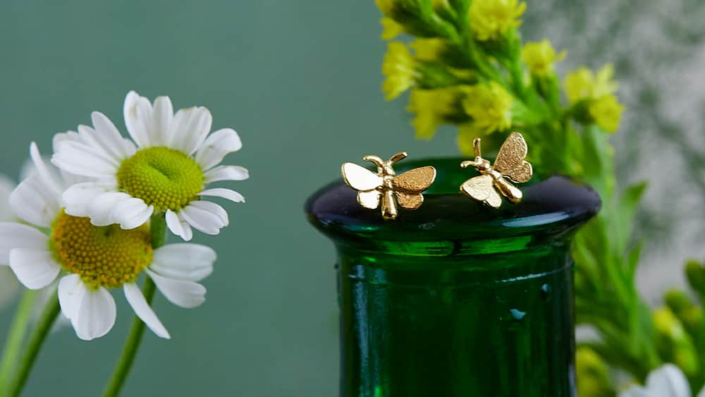 Gold tiny butterfly stud earrings on a dark green glass bottle.