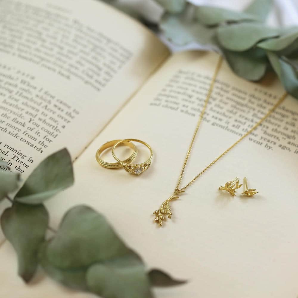 Model wearing layered gold necklaces with leaf and bird charms, a gold leaf earring, and matching rings, styled with a cream blouse in an outdoor meadow setting.